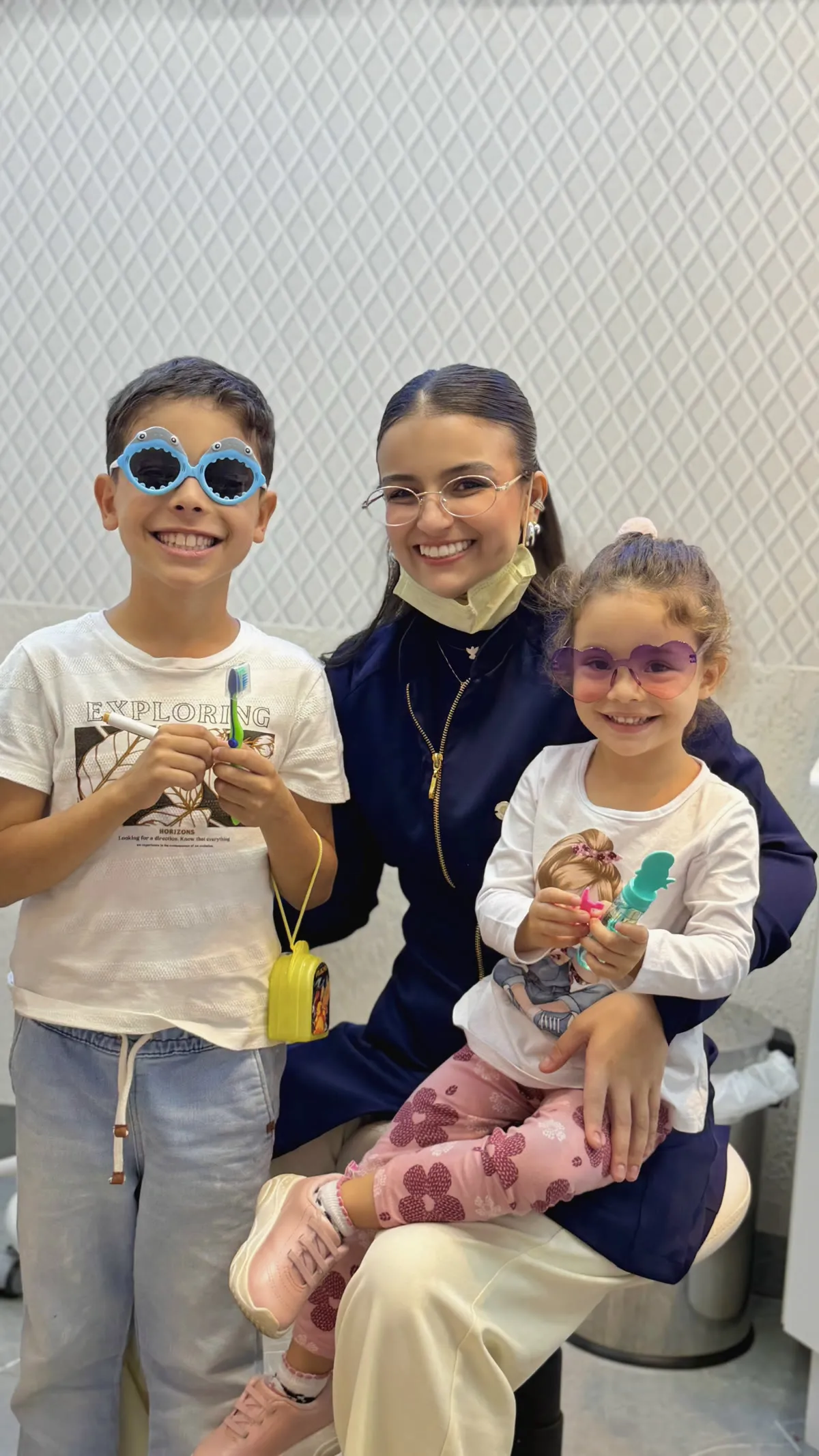 Dra. Amanda Miranda sorrindo no consultório, com duas crianças de óculos coloridos